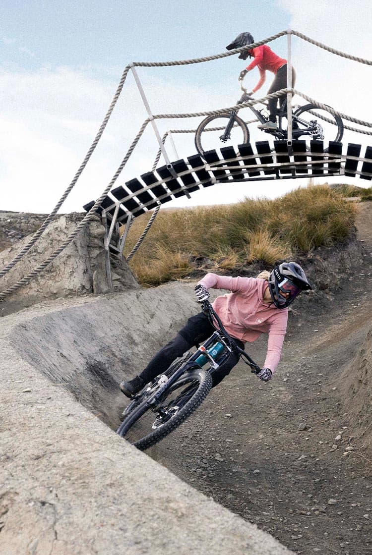 A mountain biker rides around a banked section on a Cardrona Bike Park trail as another rides a bridge over the top of them.