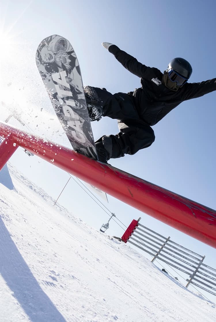 A snowboarder performing a trick while airborne over a bright red rail in a snow-covered terrain park. The rider is in mid-motion, board vertical to the ground, showcasing the artwork on the underside of the board.  There is a snowy mountain landscape in the background, on a clear blue day, along with other park features and a few spectators.