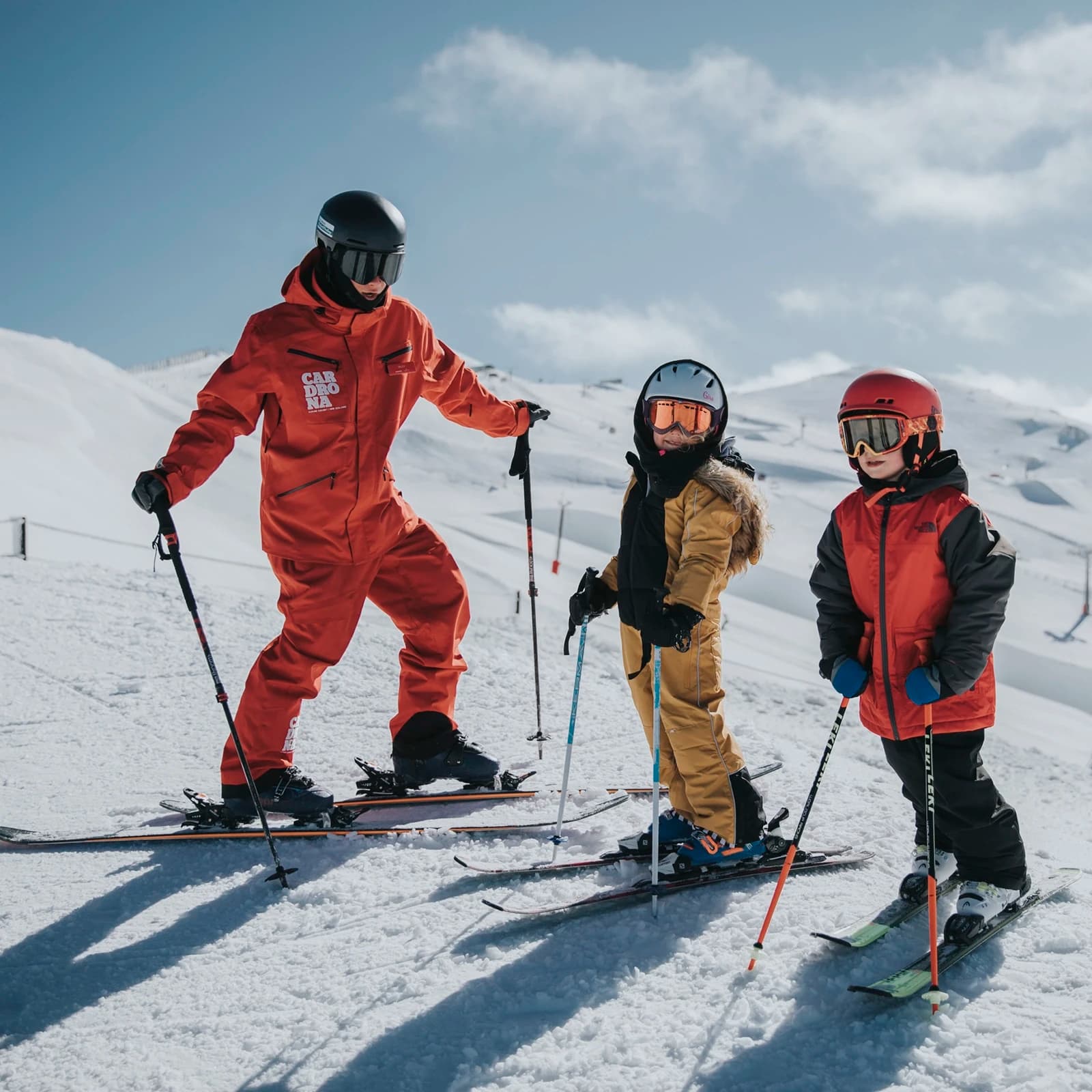 An adult ski instructor in a red snowsuit branded "Cardrona" is guiding two young children on a snowy mountain slope. The children, wearing helmets and ski gear, are standing on skis with poles in hand, listening attentively to the instructor. One child is dressed in a gold snowsuit, while the other wears a red and grey snowsuit. The snow-covered mountains stretch into the background under a bright blue sky, with ski lifts and slopes visible in the distance.