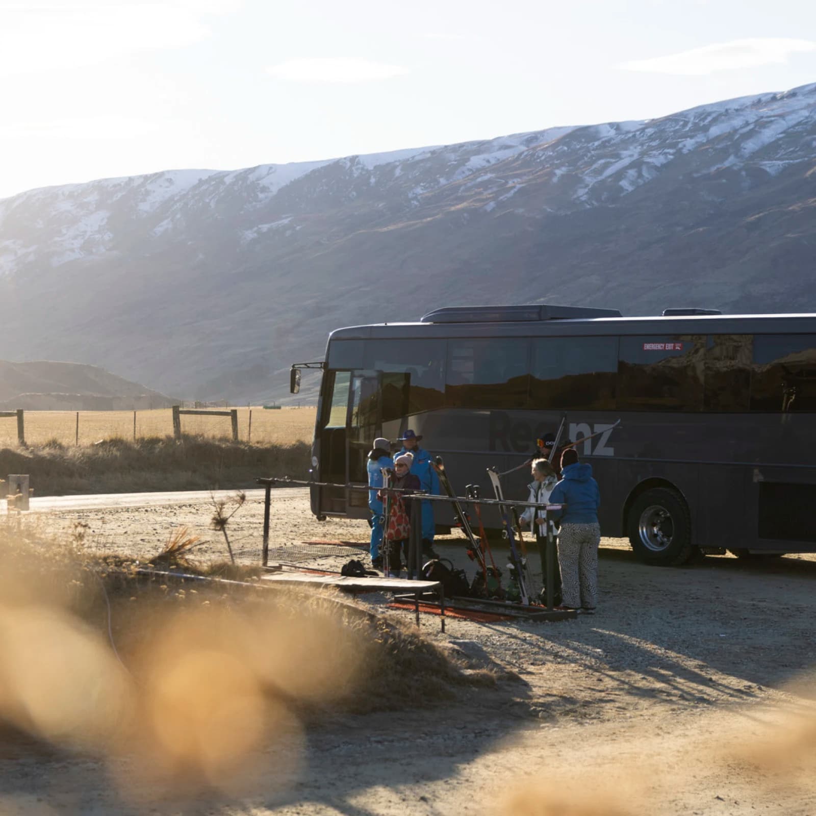 A group of people standing near a parked tour bus in a rural, mountainous area. The bus, branded "RealNZ," is dark-coloured and parked on a dirt road, there are skis and ski poles propped up nearby.