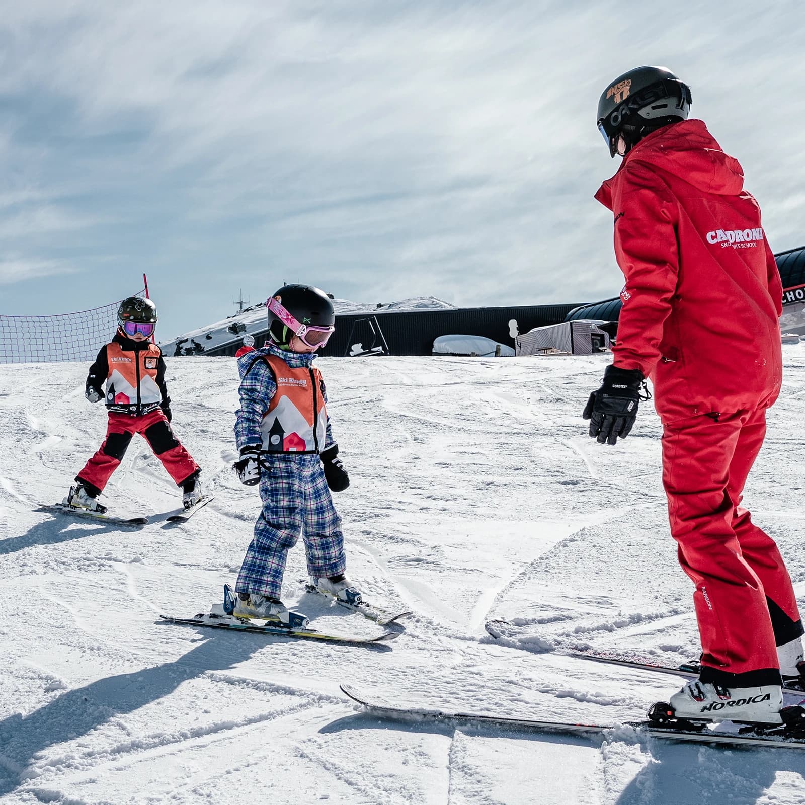 Ski instructor in red guiding two young children in orange vests as they learn to ski on a snowy slope at Cardrona, New Zealand.
