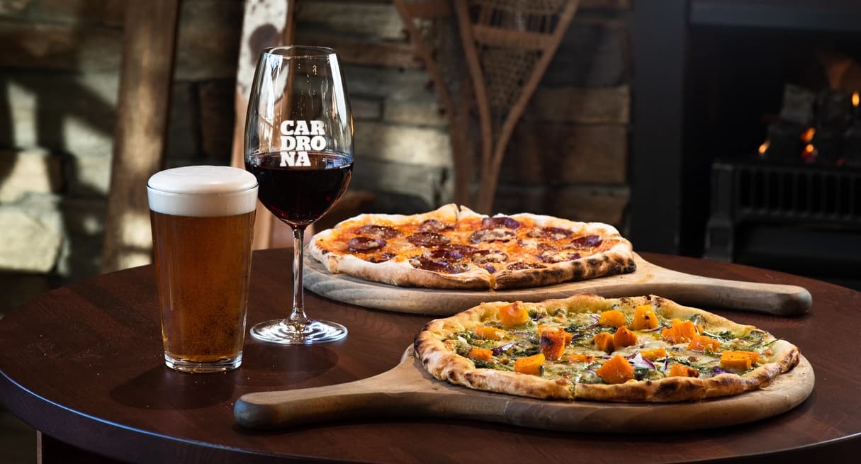 Wood-fired pizzas with a pint of beer and a glass of red wine on a table by a rustic fireplace at Cardrona.