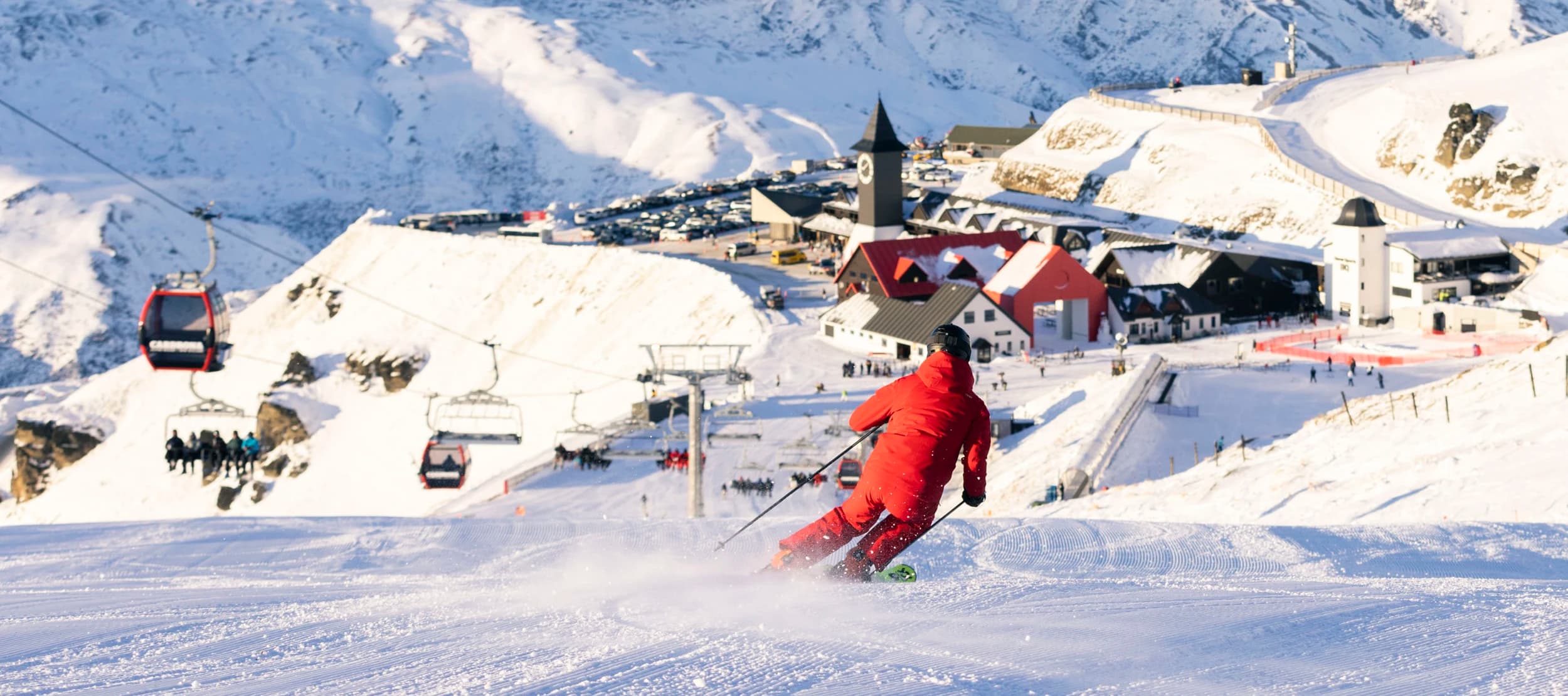 A skier dressed in red descends a freshly groomed slope at Cardrona, with the village, gondola, and chairlift visible in the background, all set against the snow-covered mountains.