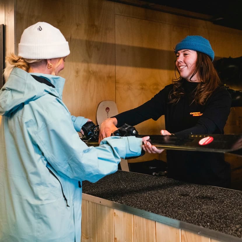 Two people inside a ski rental shop. On the left, a customer in a light blue ski jacket and white beanie is receiving a pair of skis from a staff member. The staff member, smiling, is wearing a blue beanie and a black shirt. They are standing at a wooden counter, with skis and equipment visible in the background.