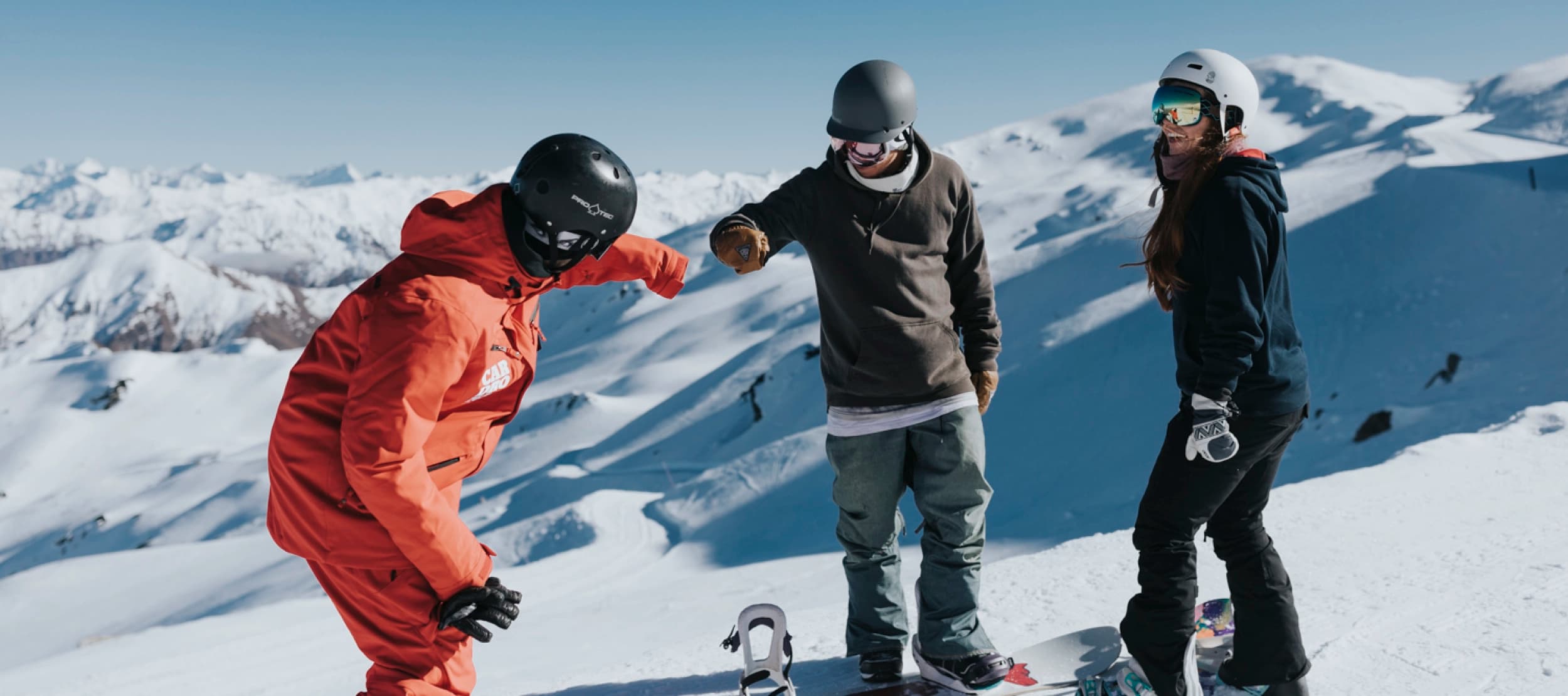 Three snowboarders stand on a snowy mountain. They are casually dressed in snow gear, including helmets and goggles, with snowboards at their feet. One snowboarder in a red suit is fist-bumping another person dressed in a grey jacket and green pants, while a third person in a navy blue jacket and long brown hair watches, smiling.