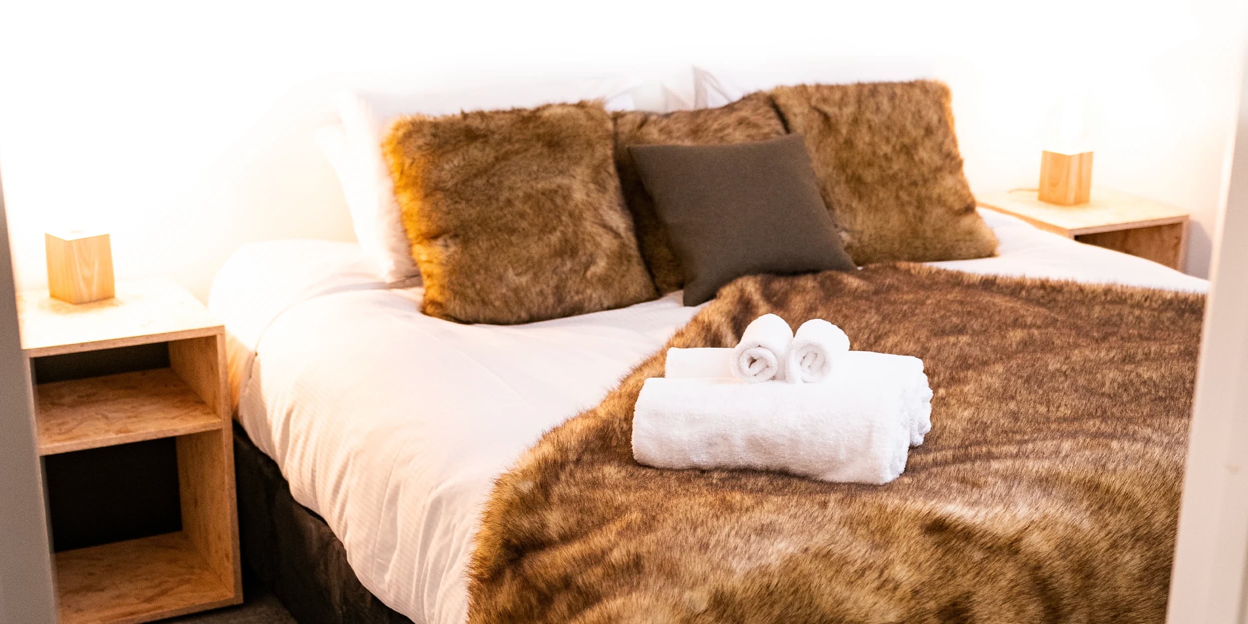 A cosy bed with white sheets and brown fur throw and cushions.