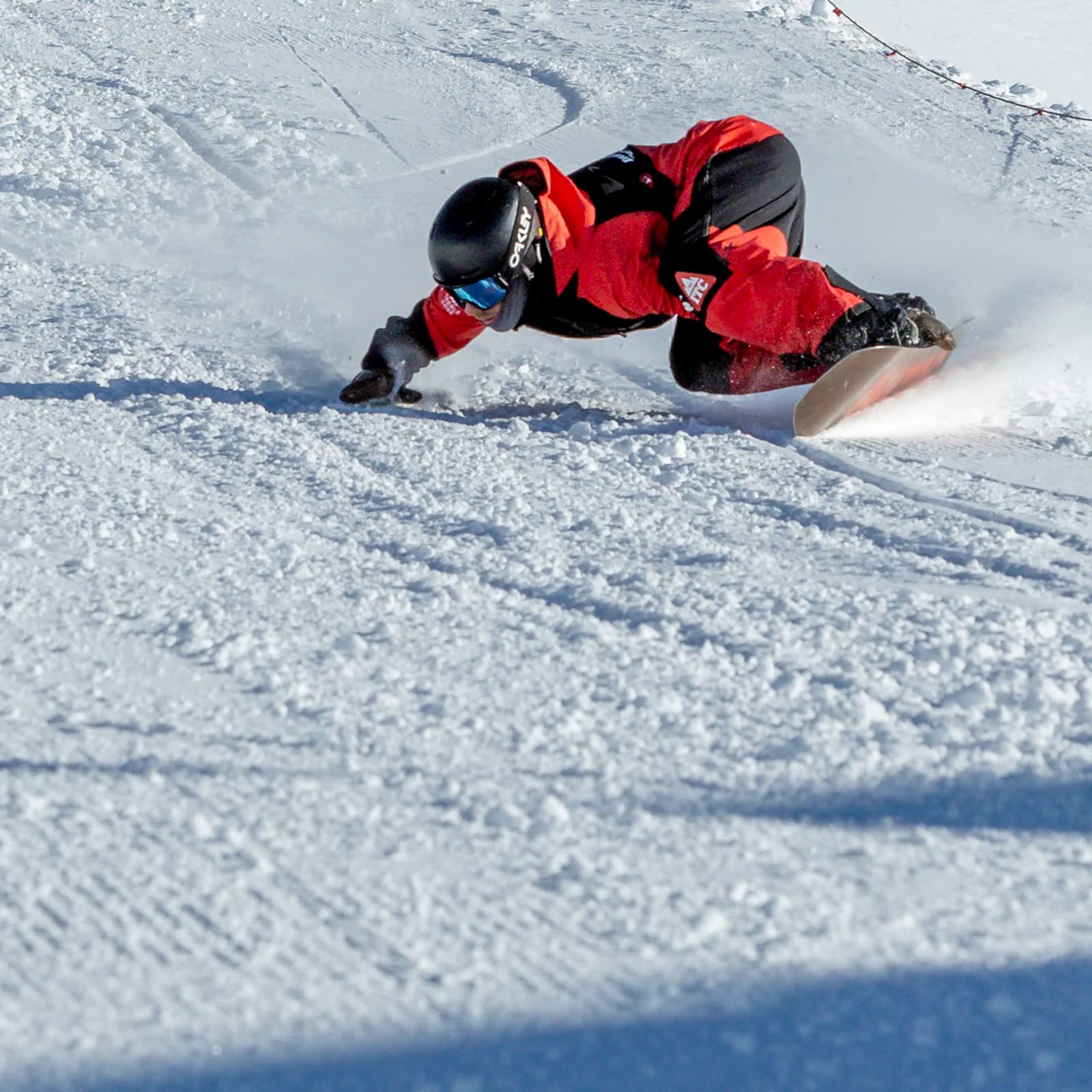 A snowboarder, wearing a black helmet and red jacket, leans sharply into the snow as they carve through a slope, creating a spray of snow in their wake.