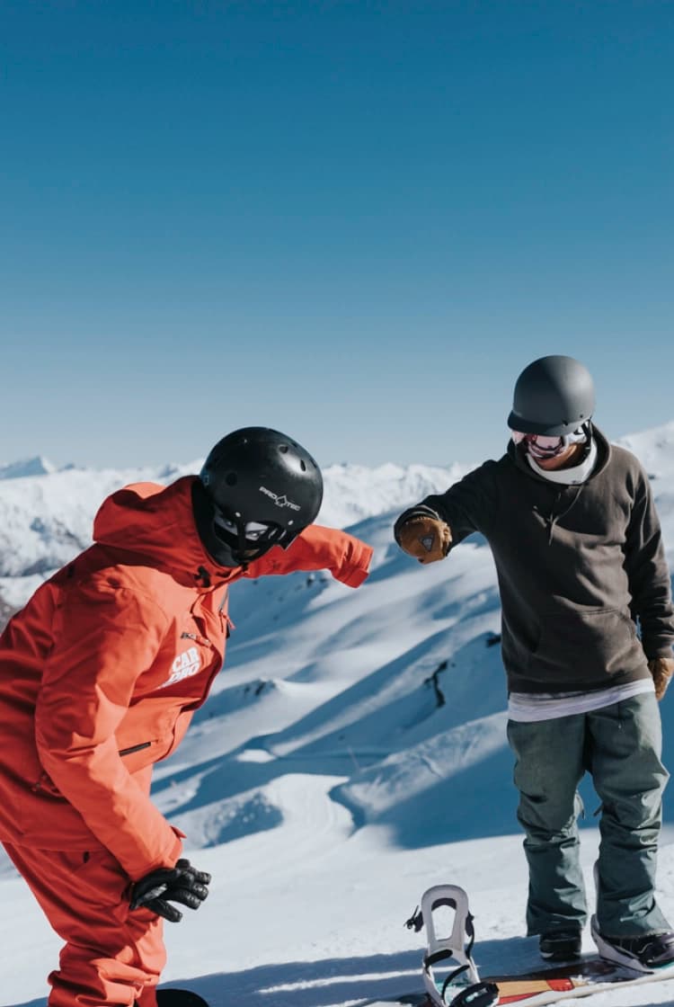 Two snowboarders stand on a snowy mountain. They are casually dressed in snow gear, including helmets and goggles, with snowboards at their feet. One snowboarder in a red suit is fist-bumping the other, dressed in a grey jacket and green pant.