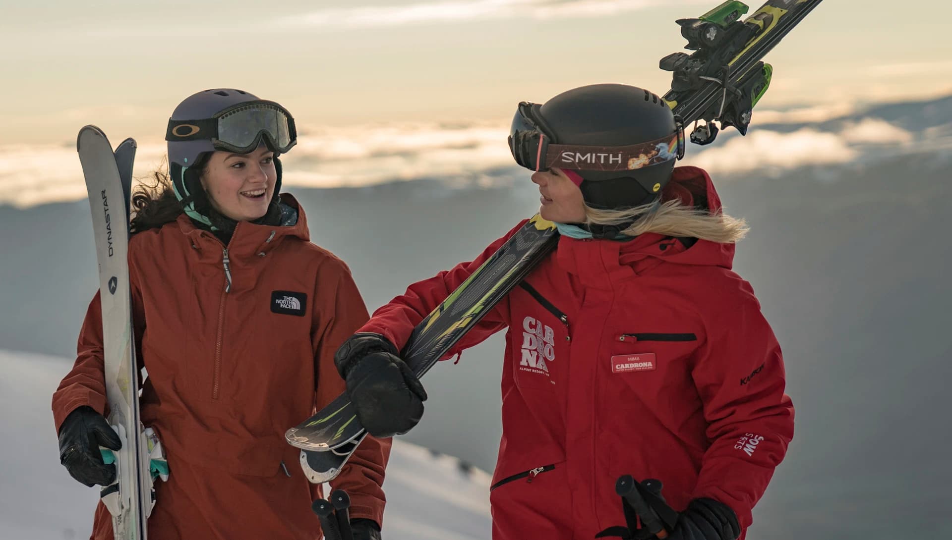 Two women are standing on a snowy mountain engaged in conversation. Both women are wearing ski gear and carrying skis on their shoulders. One of the women is dressed in a red Cardrona Alpine Resort jacket.
