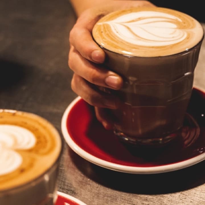 Two lattes with latte art served in glass cups on red saucers, one being held by a hand.