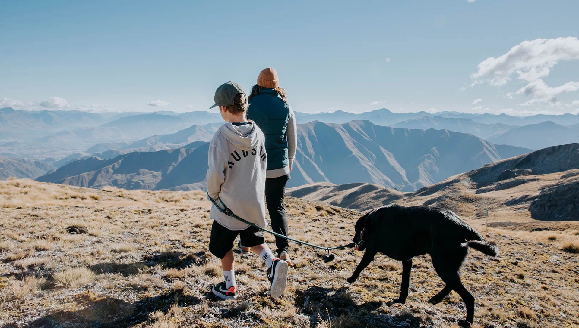 A woman, child and a black dog walk along mountainous terrain on a clear sunny day. The background features a panoramic view of softly lit mountain ranges.