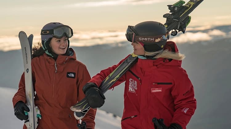 Two women are standing on a snowy mountain engaged in conversation. Both women are wearing ski gear and carrying skis on their shoulders. One of the women is dressed in a red Cardrona Alpine Resort jacket.