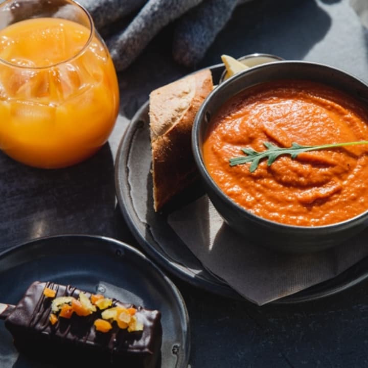 Tomato soup with bread, a glass of orange juice, and a chocolate dessert on a dark table in sunlight.