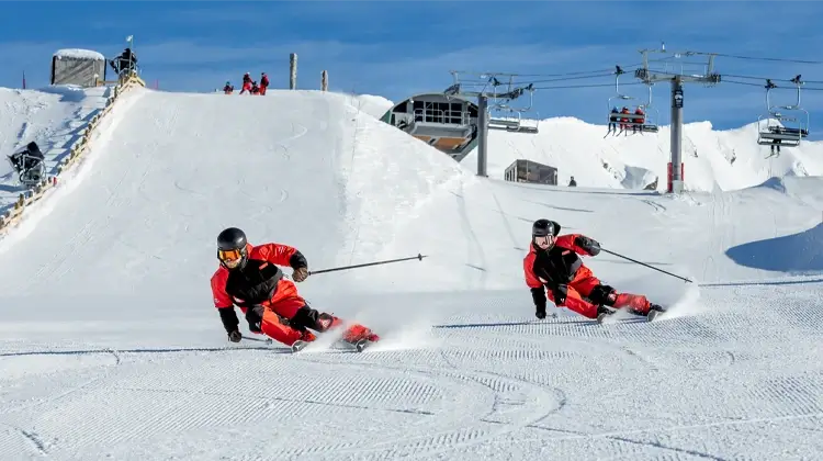 Two skiers training at Cardrona's Instructor Training Centre, carving precisely down a groomed slope beneath chairlifts on a clear, sunny day.