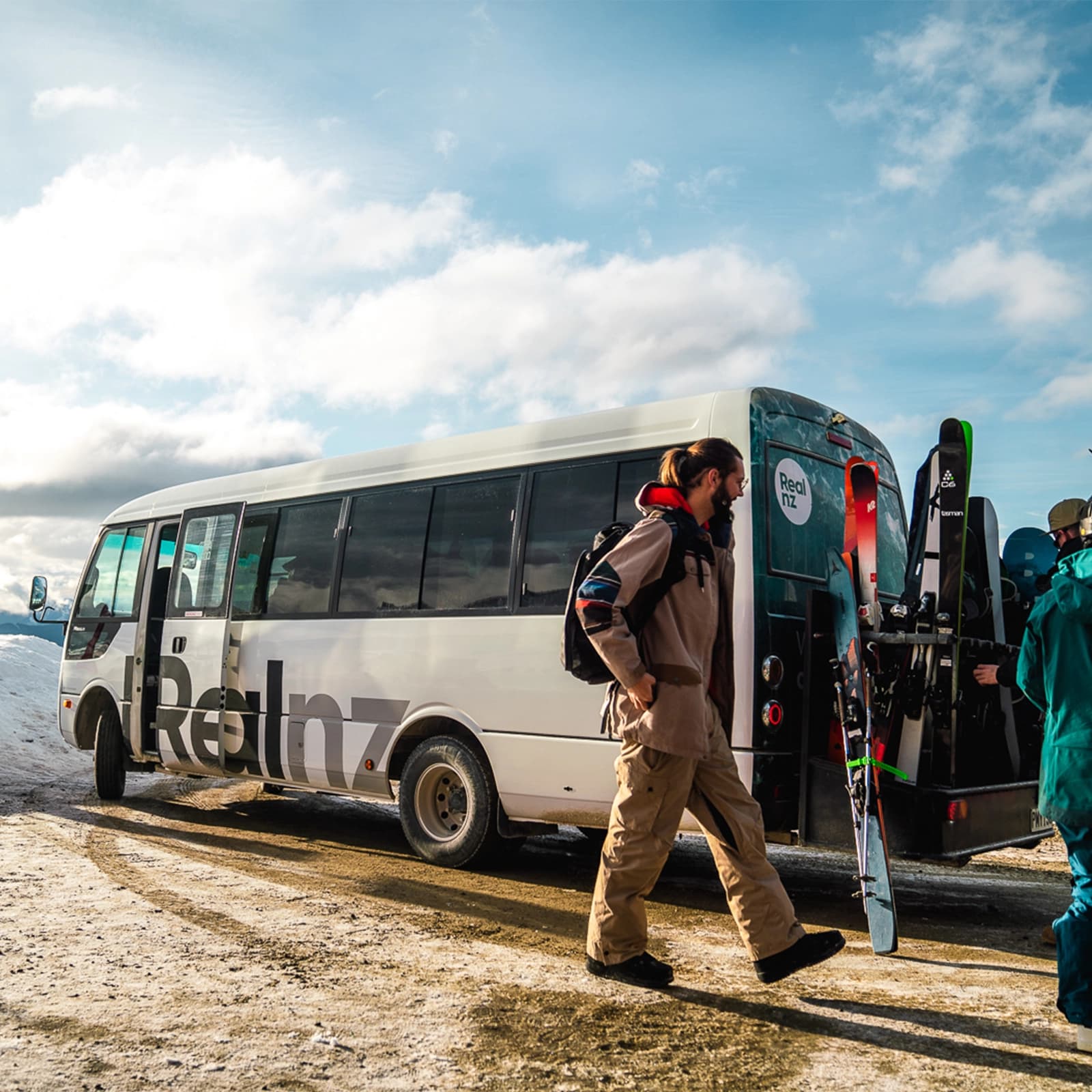 A group of skiers prepares their gear next to a white bus in a snowy mountain setting, ready for transport to their destination. Clear skies and bright sunlight overhead.