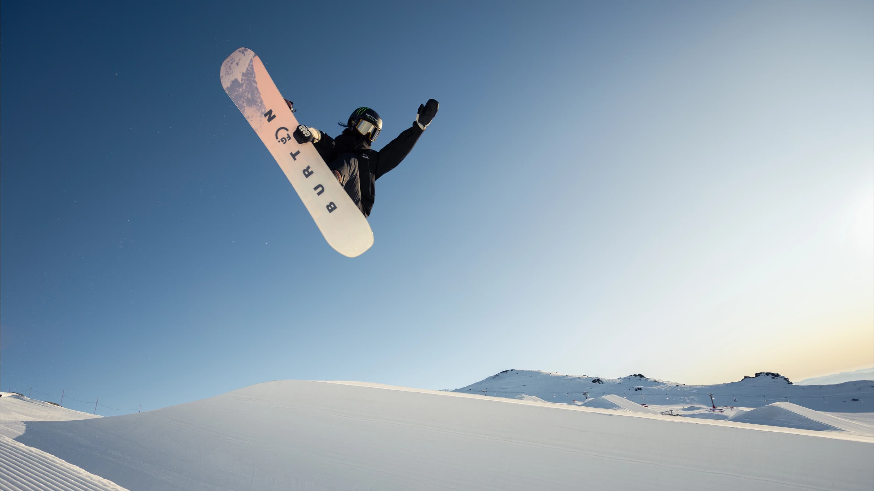 Snowboarder catching big air in the Cardrona mini pipe against a clear blue sky.