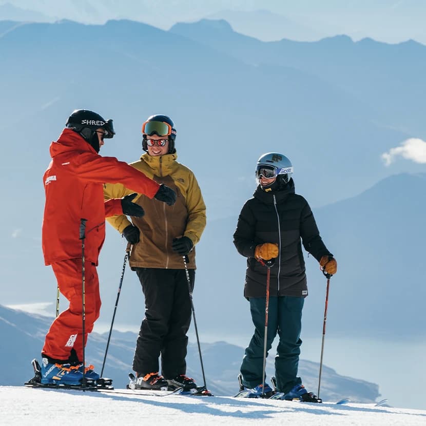 A ski instructor and two skiers stand on a snowy mountain slope, engaging in a lesson. The instructor, dressed in a bright red Cardrona jacket and pants, is gesturing and providing guidance to the two skiers.