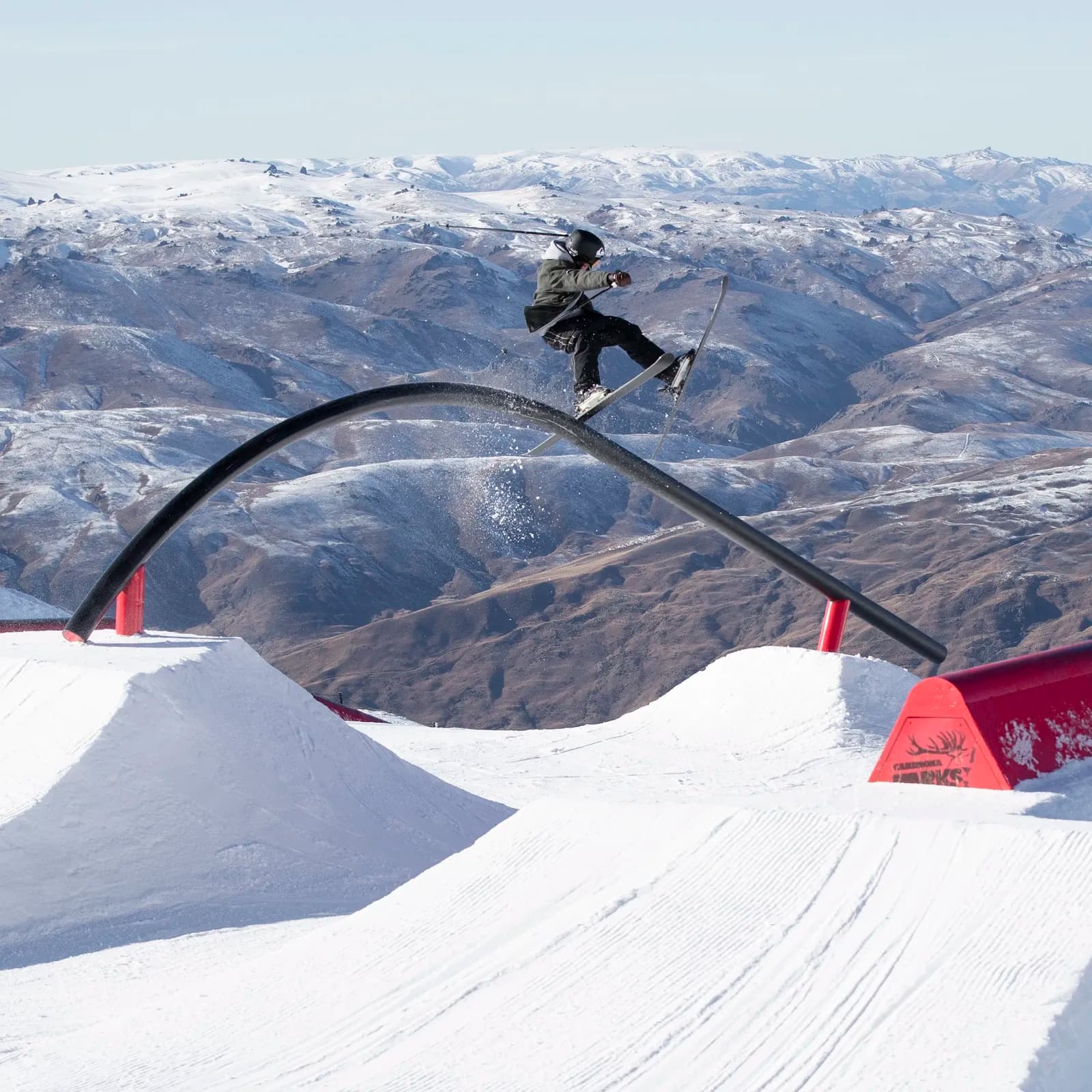 A skier performs a trick on a black rail in a snow-covered terrain park. The background shows rolling mountains dusted in snow on a clear blue day.