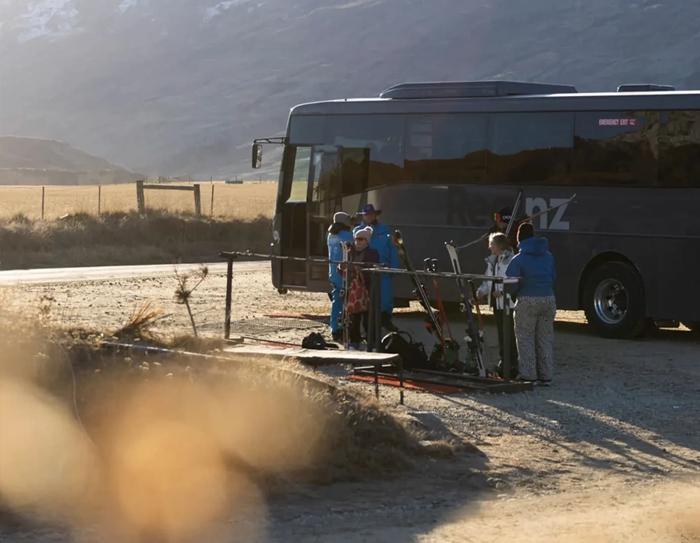 Skiers preparing equipment beside a shuttle bus on a gravel area.
