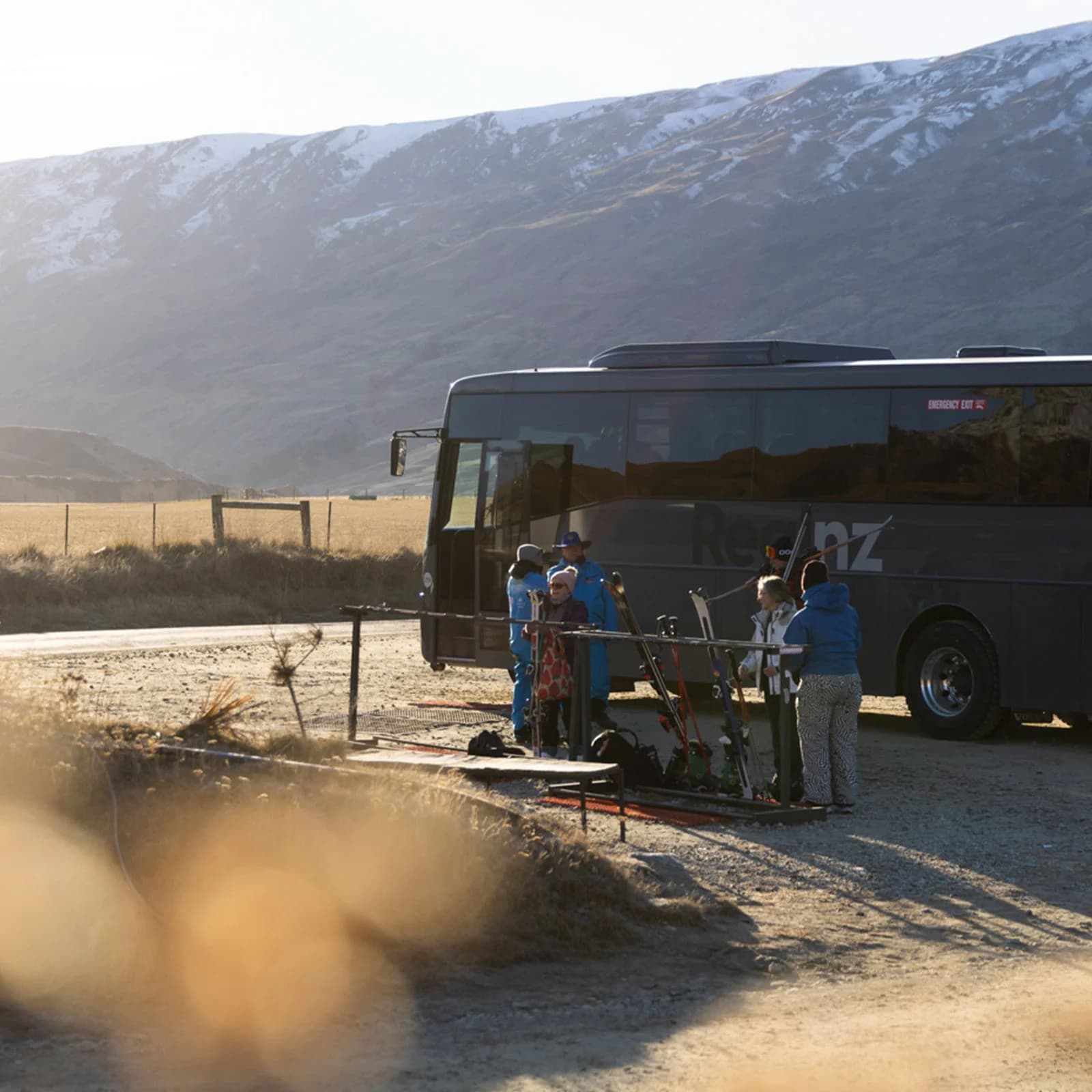 A group of skiers gathers near a parked tour bus in a mountainous, sunlit valley. They organise their gear, preparing for a day on the slopes. Snow-dusted hills rise in the background, creating a peaceful outdoor scene.