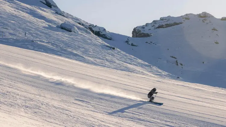 A skier glides down a wide, smooth, snow-covered slope under bright sunlight. The skier's shadow stretches out behind them as they carve gracefully, leaving a thin trail of snow dust in their wake. The surrounding mountain landscape is bathed in soft, golden light, with rocky, snow-dusted peaks rising in the background.