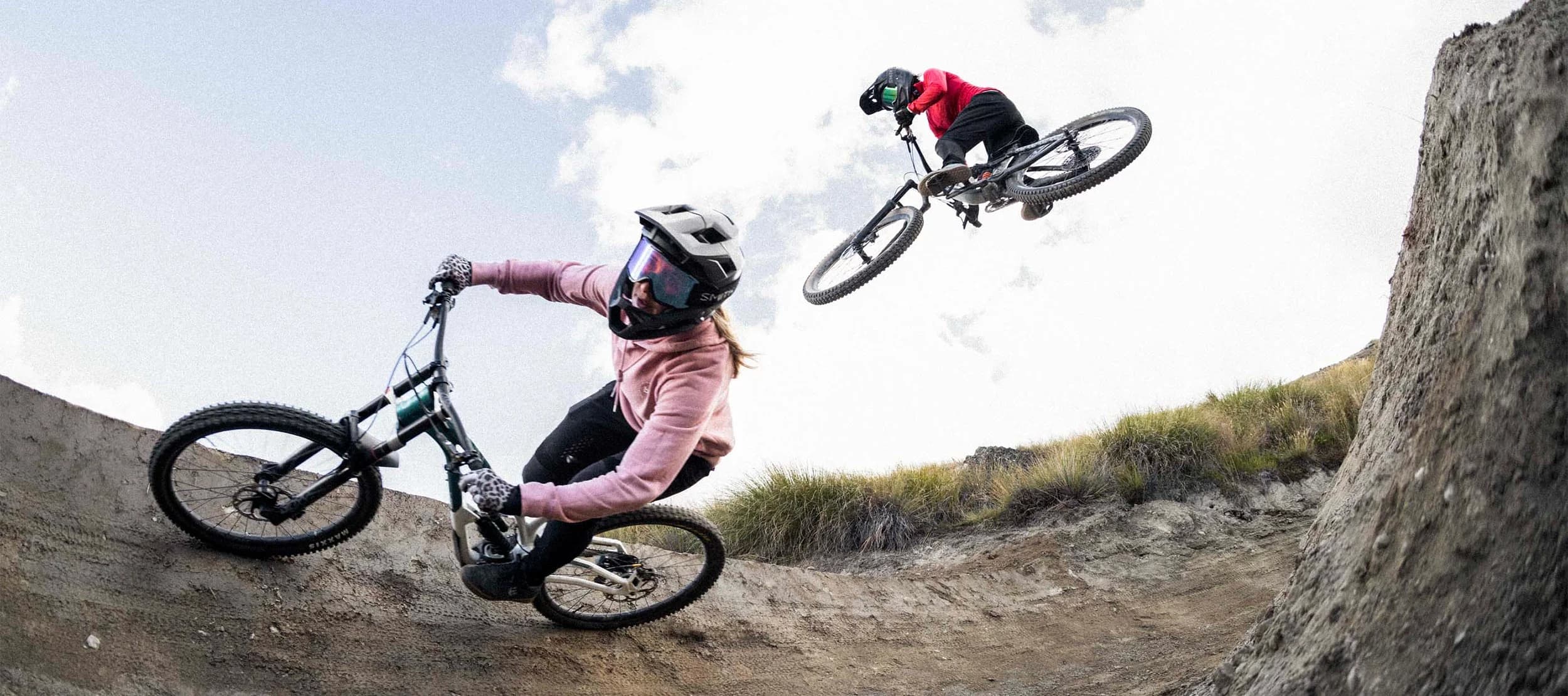 A mountain biker rides around a banked section on a Cardrona Bike Park trail as another jumps over the top of them.