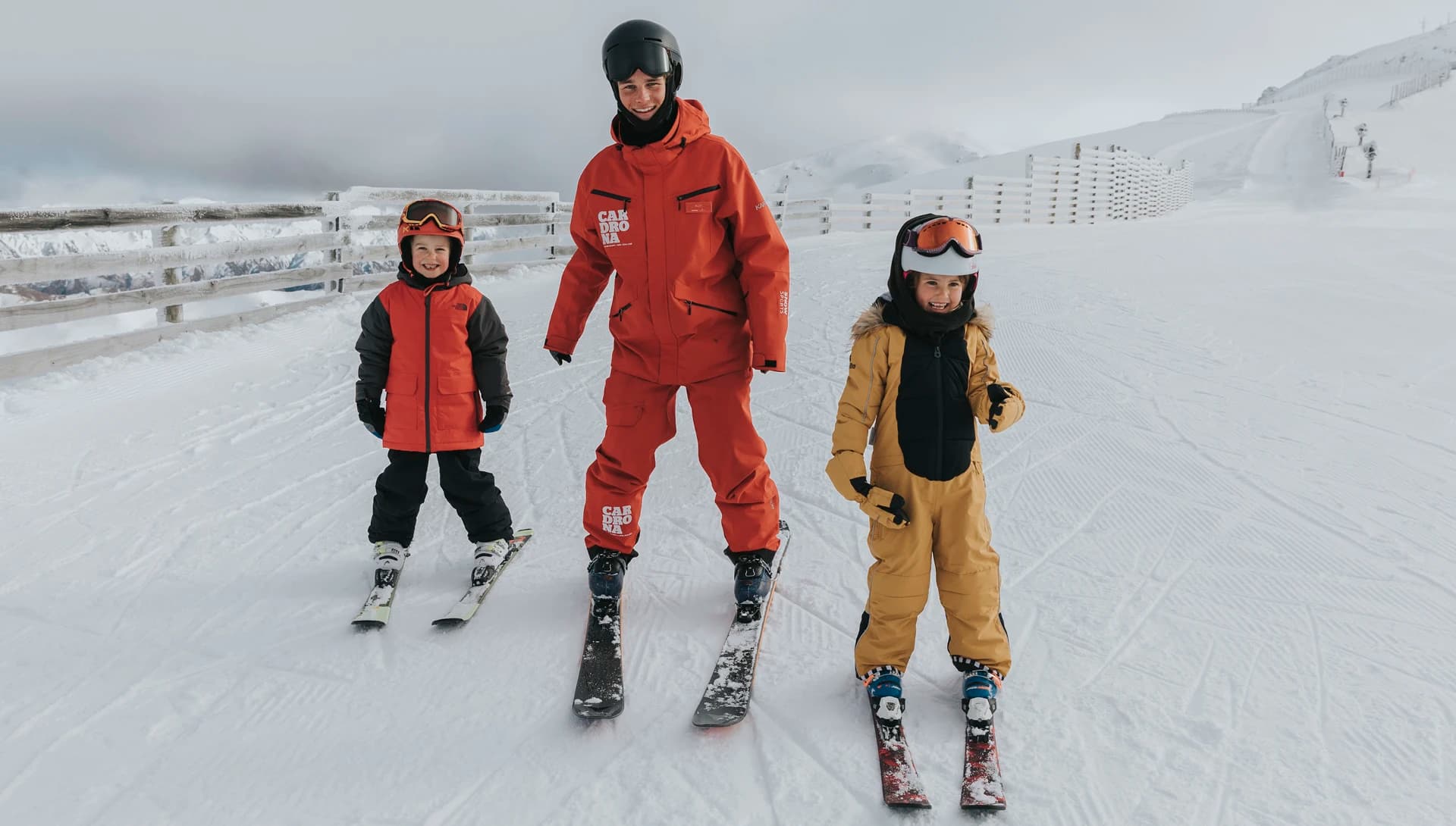 A ski instructor and two young children stand together on a snowy mountain, ready for a skiing session. The instructor, dressed in a bright red snowsuit branded "Cardrona", smiles while standing on skis. The two children, one wearing a red and black snowsuit and the other in a gold snowsuit, also have helmets and goggles. They are all positioned on a snow-covered platform with a wooden fence behind them, and the mountain's ski slopes can be seen in the distance under a cloudy sky.