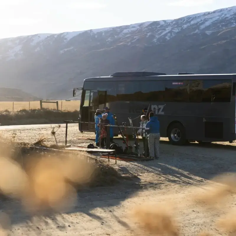 A group of skiers wait to board a RealNZ bus as the bottom of a snowy mountain.