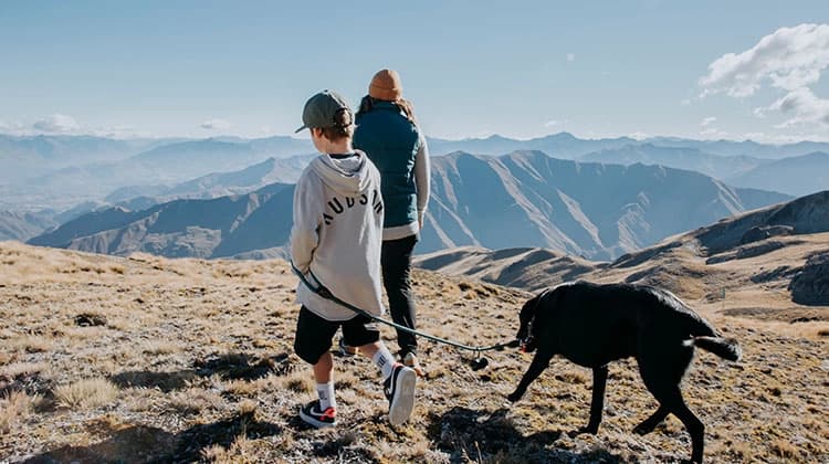 A woman, child and a black dog walk along mountainous terrain on a clear sunny day. The background features a panoramic view of softly lit mountain ranges.