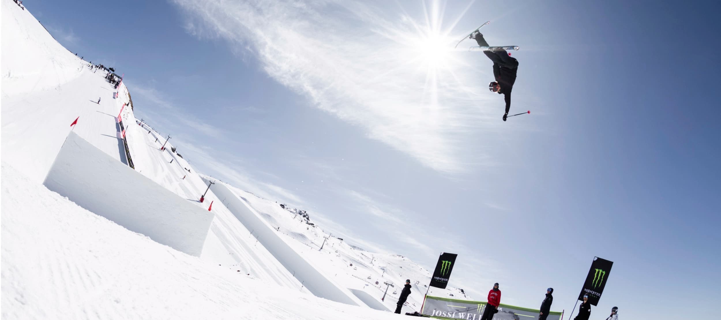 A skier is performing an aerial trick high above a snowy slope, captured mid-flip with one arm extended, while the bright sun shines behind them in a clear blue sky. Below, several people watch from the side of the park, near a branded banner and flags. The snow park features ramps and jumps, with other skiers visible in the distance.