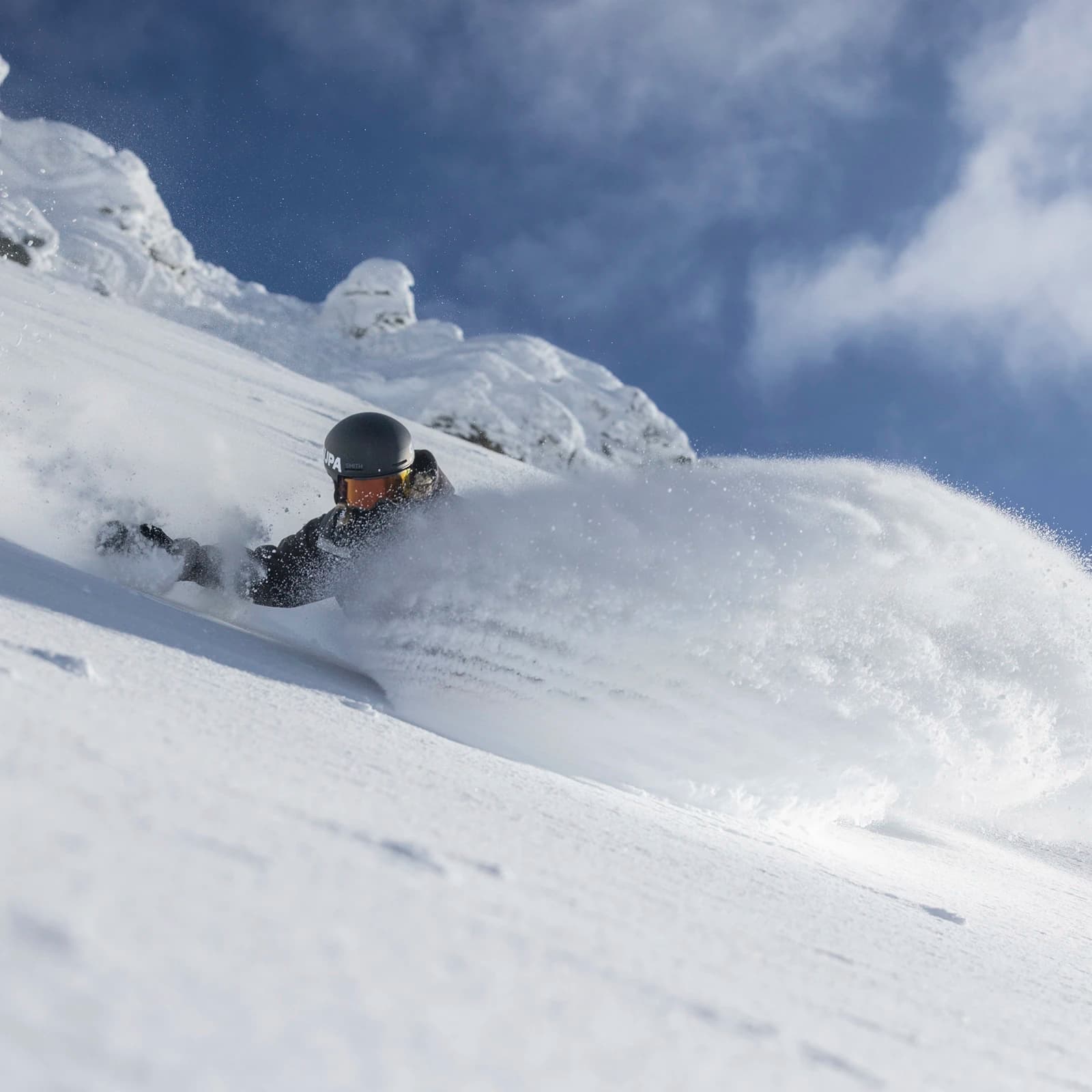 A snowboarder carves through deep, fresh powder on a snowy mountain slope. Their helmet and goggles reflect the bright sunlight, while a huge spray of snow arcs behind them, capturing the speed and energy of the descent. The background features rugged, snow-covered peaks against a clear, blue sky.