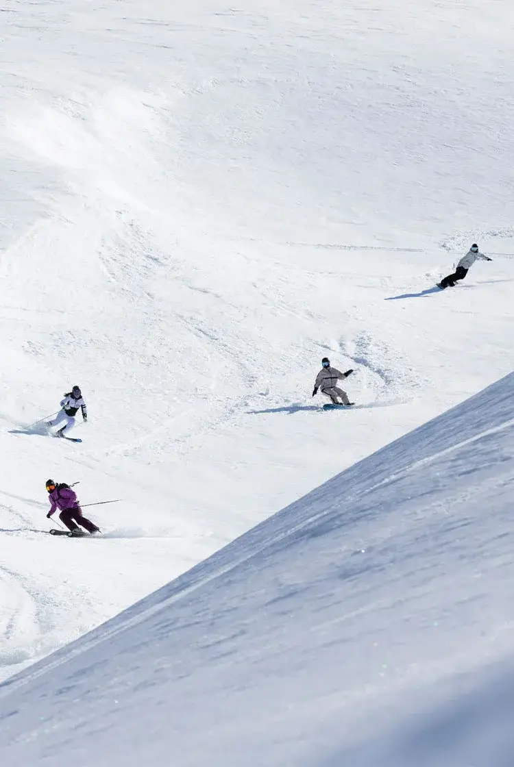 Group of skiers and snowboarders carving down a snowy slope under a clear sky.