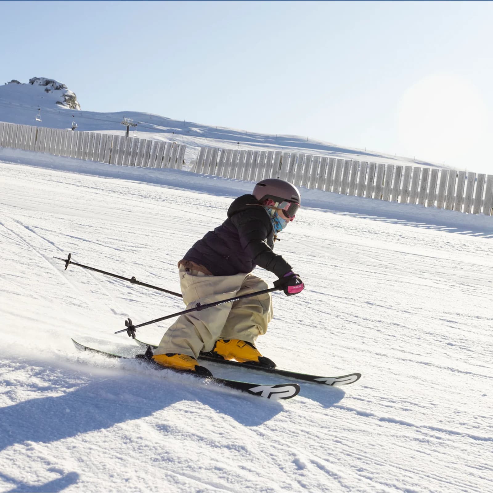 A young skier carves down a sunlit slope, leaning into the turn with focus and control. Dressed in winter gear and helmet, they glide smoothly across the snow with ski poles in hand. A fence and snow-covered landscape stretch out in the background under a clear sky.