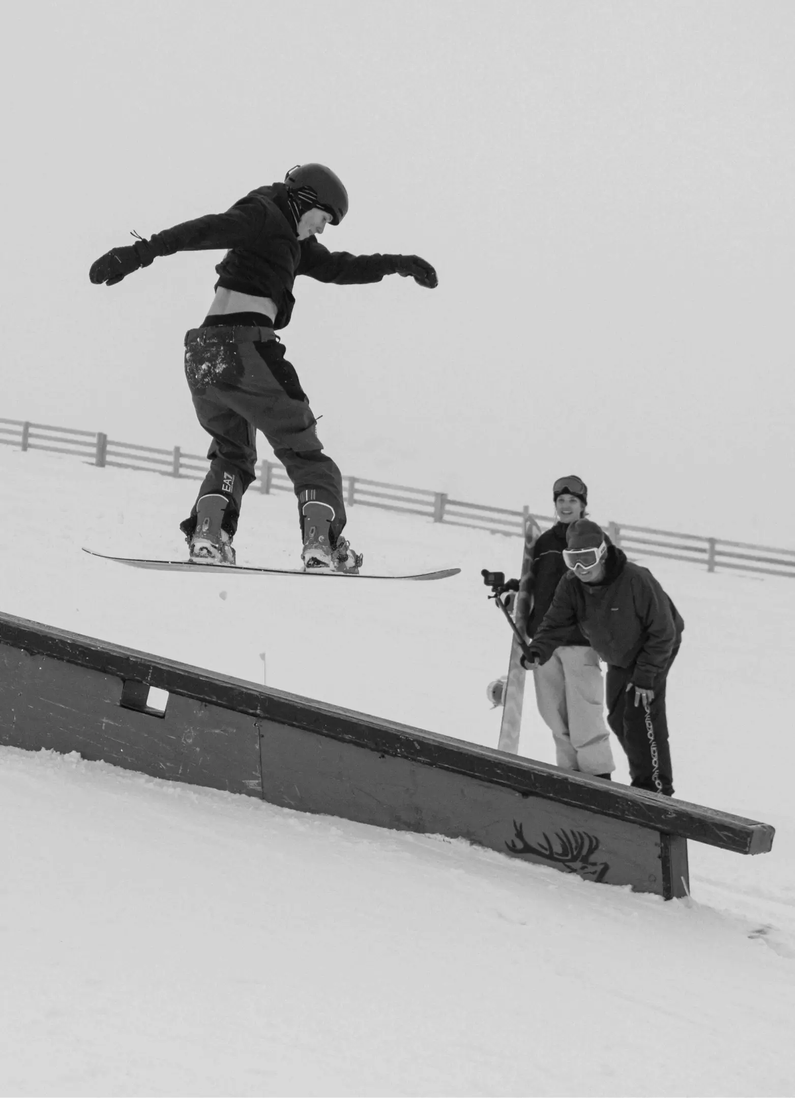 Snowboarder performs a jump onto a rail in a terrain park while two people watch and smile in the background on a snowy mountain.