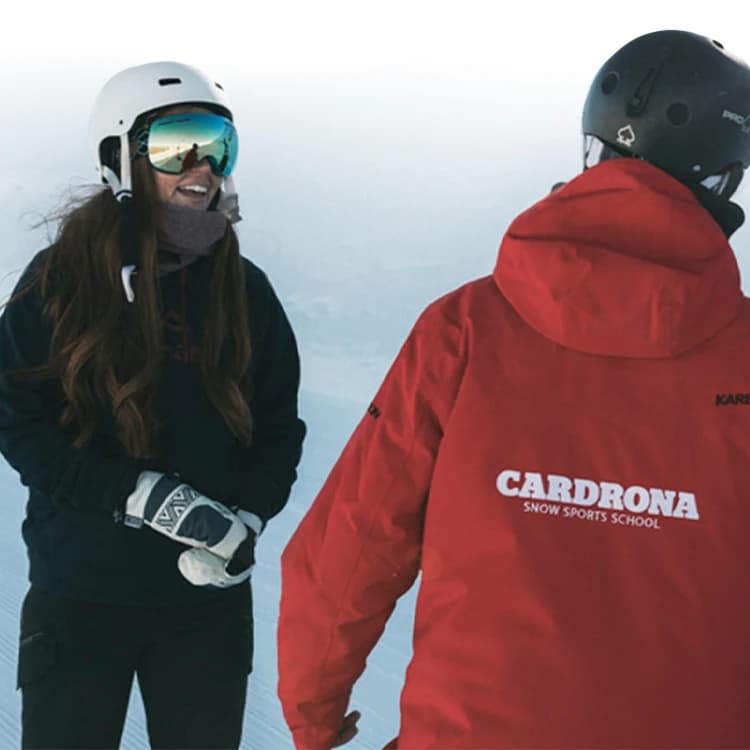 Two skiers or snowboarders standing together on a snowy mountain slope. Both are wearing ski gear, one is facing away from the camera and wearing a red jacket with "CARDRONA SNOW SPORTS SCHOOL" printed on the back.