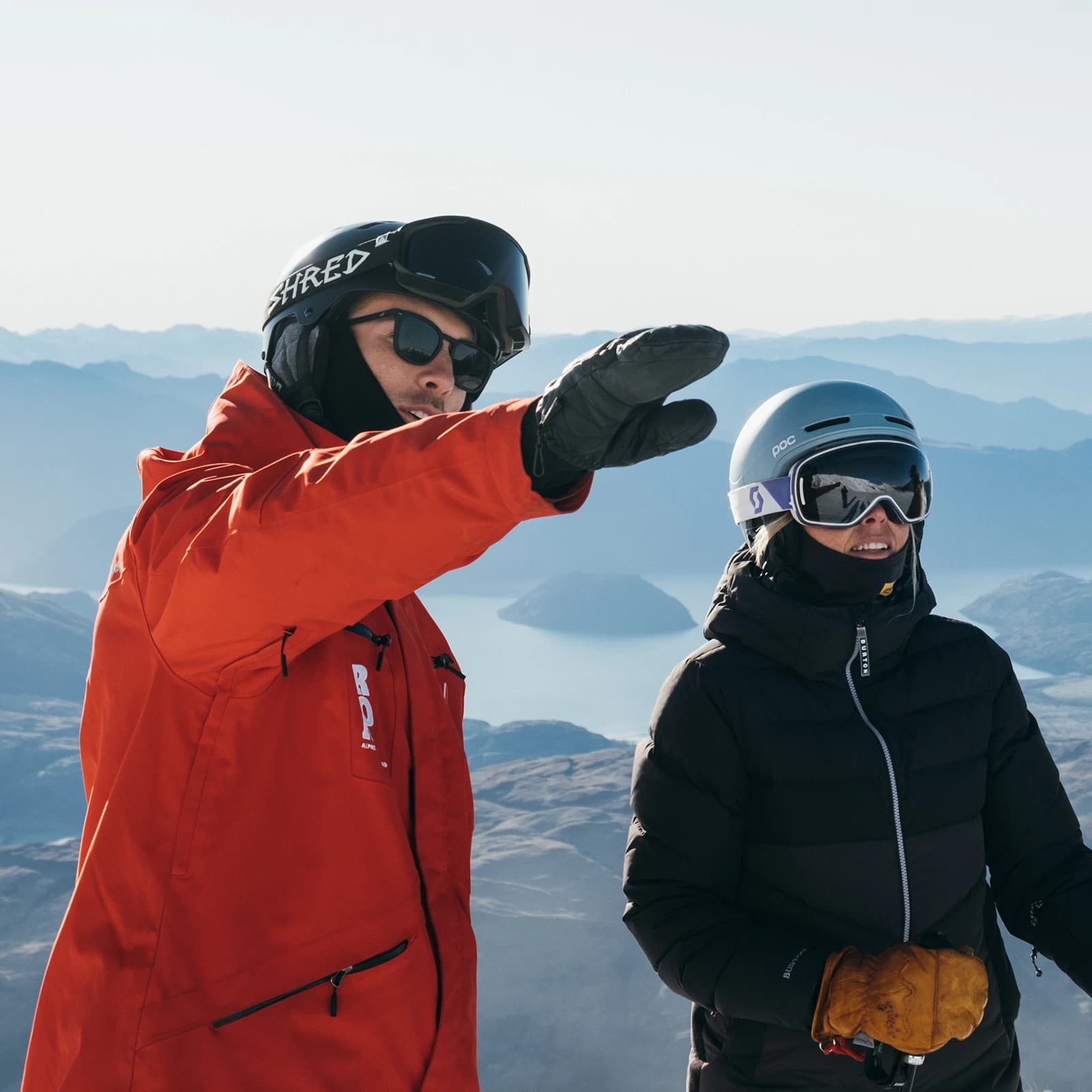 Two skiers in winter gear stand on a mountain; one in a red jacket points out over the landscape while the other looks on, with a scenic lake and peaks in the background.