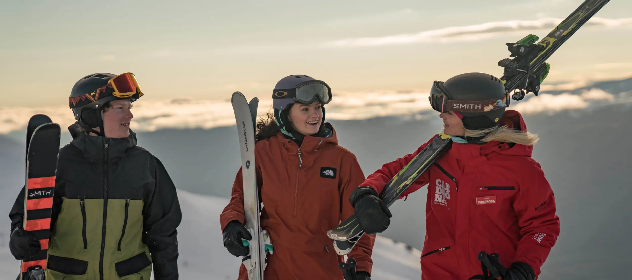An instructor and two learners dressed in winter gear chat at Cardrona Alpine Resort, carrying skis with a sunset and mountain backdrop, ready for a day on the slopes.