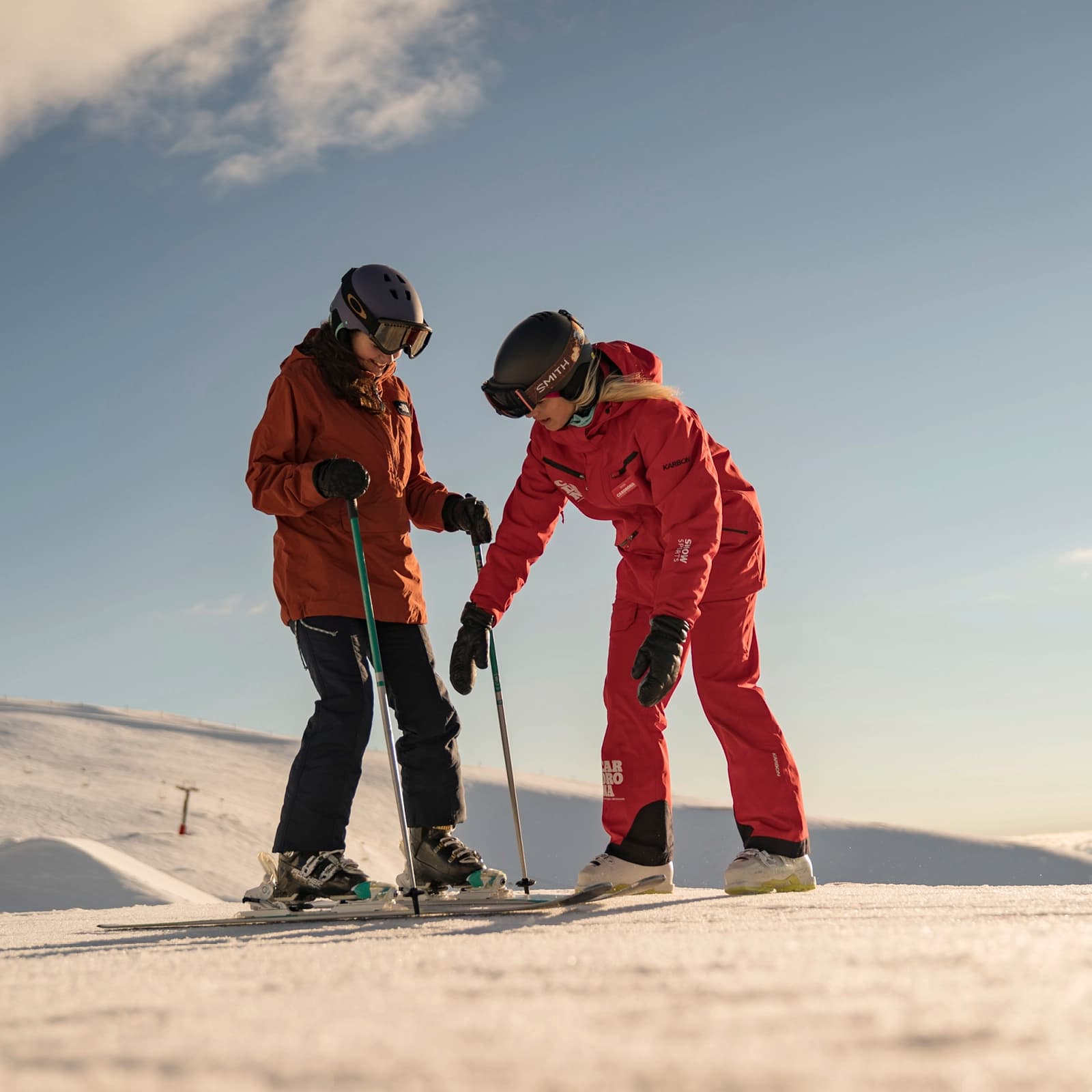 Two skiers on a gentle snowy slope under a clear sky. One person, dressed in red ski gear with a helmet and goggles, appears to be giving instructions or helping the other skier. The second skier, in an orange jacket and dark pants, is standing attentively with their ski poles. Both are equipped with skis and winter gear, suggesting a ski lesson or guidance for a beginner.