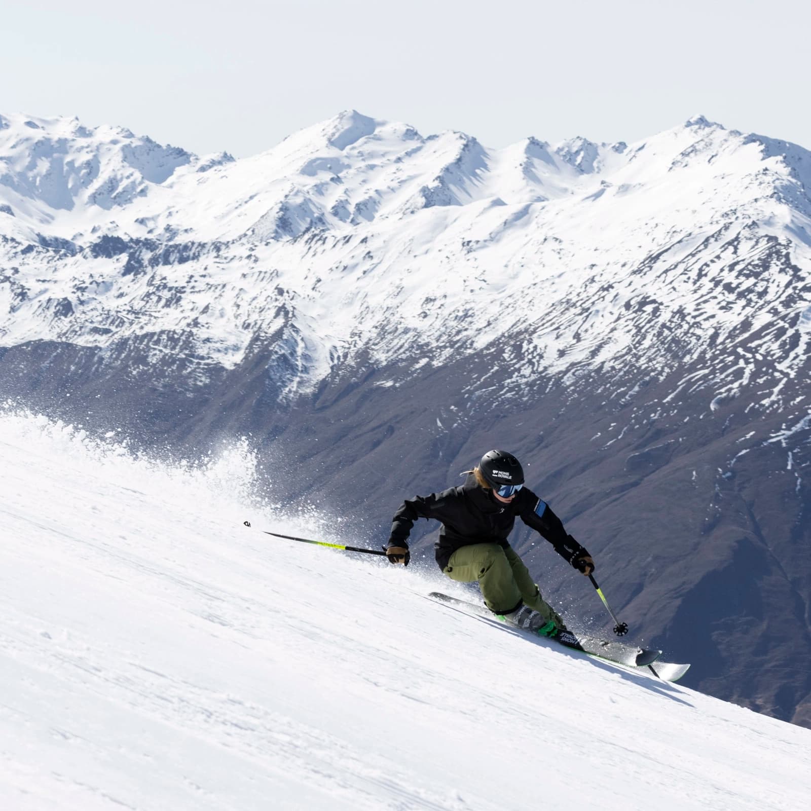 Skier in action, speeding down a snowy mountain slope against a backdrop of stunning snow-covered peaks of the New Zealand Southern Alps. The skier, dressed in a black jacket, green pants, and wearing a helmet and goggles, is carving through the snow, creating a spray of powder in their wake. The clear sky and expansive mountain range in the background add to the feeling of adventure and freedom in the winter landscape.