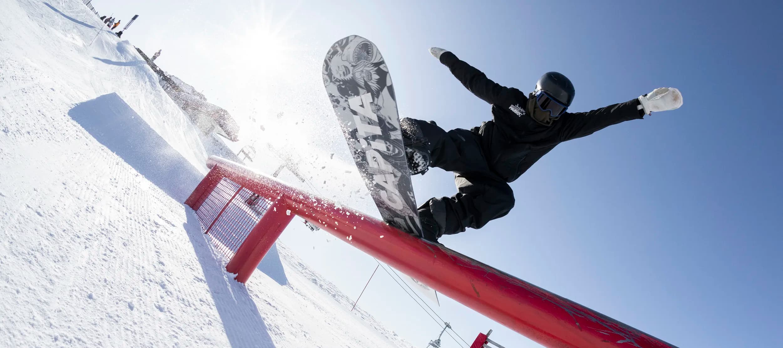 A snowboarder performing a trick while airborne over a bright red rail in a snow-covered terrain park. The rider is in mid-motion, board vertical to the ground, showcasing the artwork on the underside of the board.  There is a snowy mountain landscape in the background, on a clear blue day, along with other park features and a few spectators.