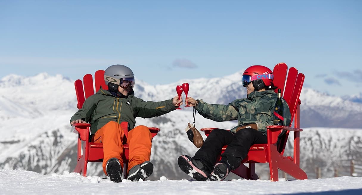 Two skiers toast on a snowy mountaintop at Cardrona in red chairs, enjoying breathtaking alpine views and a celebratory moment.
