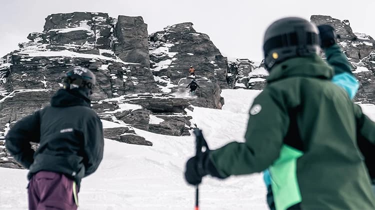 Two skiers in the foreground watch a third skier performing a jump from a rocky outcrop in a snowy landscape. The skier in the air is mid-jump.