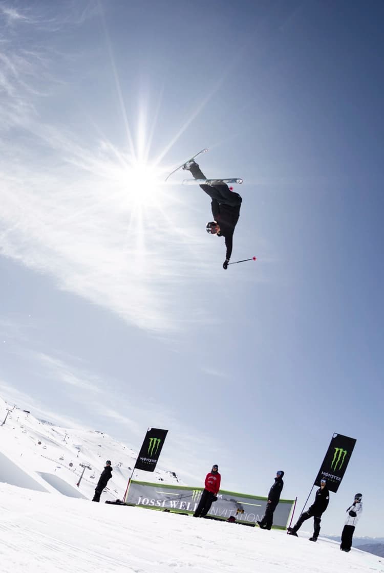 A skier is performing an aerial trick high above a snowy slope, captured mid-flip with one arm extended, while the bright sun shines behind them in a clear blue sky. Below, several people watch from the side of the park, near a branded banner and flags. The snow park features ramps and jumps, with other skiers visible in the distance.