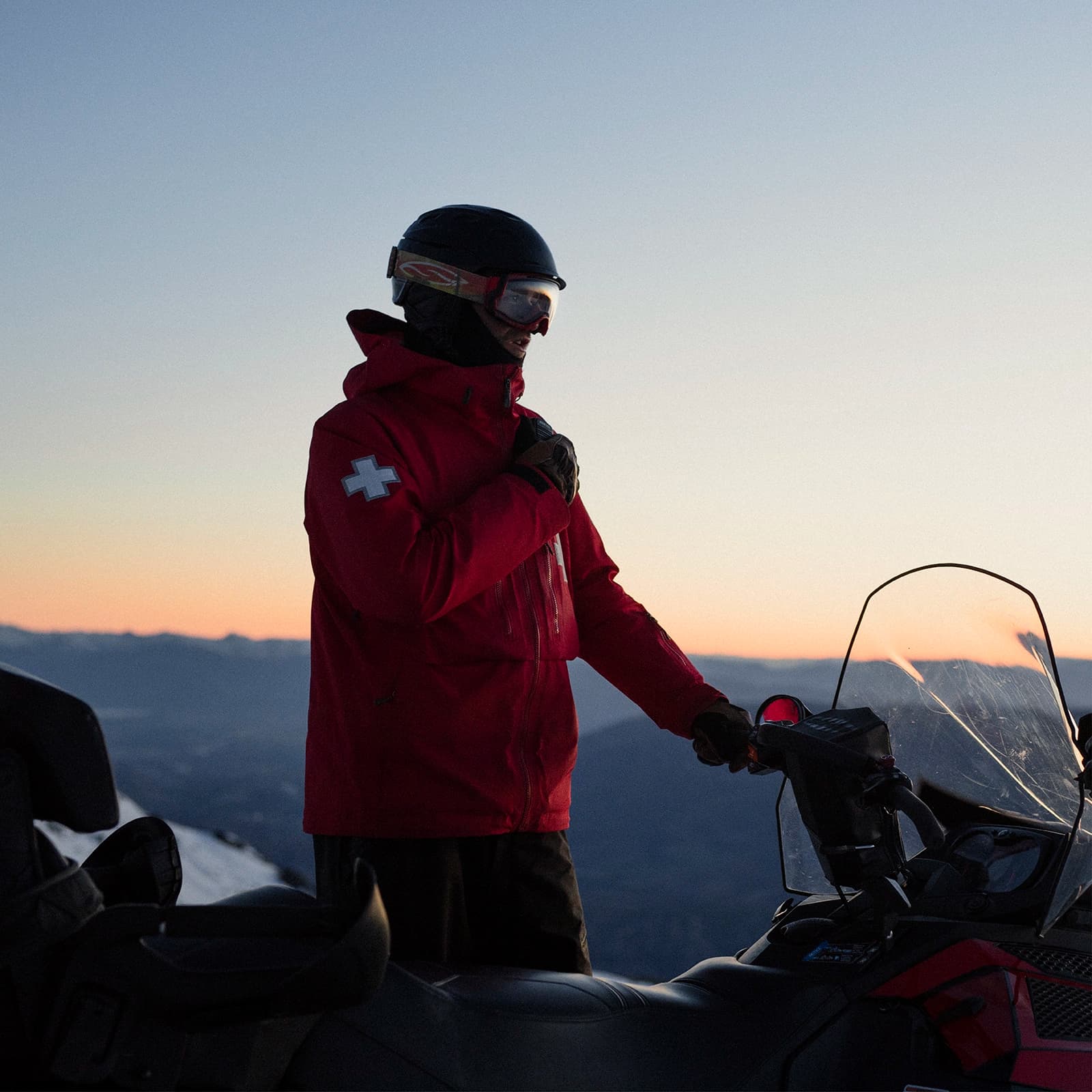 A ski patroller standing next to a snowmobile at dusk or dawn. The patroller is wearing a red jacket with a white cross on the sleeve, a helmet, and ski goggles. The scene is backlit by a soft, fading light, with mountains and a clear sky in the background, creating a calm and serene atmosphere. The patroller appears to be in a moment of preparation or communication, possibly adjusting their gear or radio.
