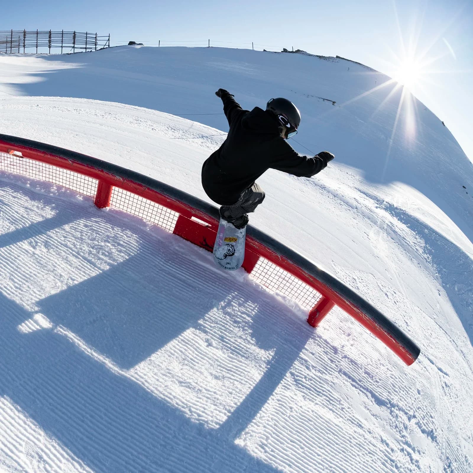 A snowboarder performing a trick over a bright red rail in a snow-covered terrain park. There is a snowy mountain landscape in the background on a clear blue day with the sun shining from behind the mountain.
