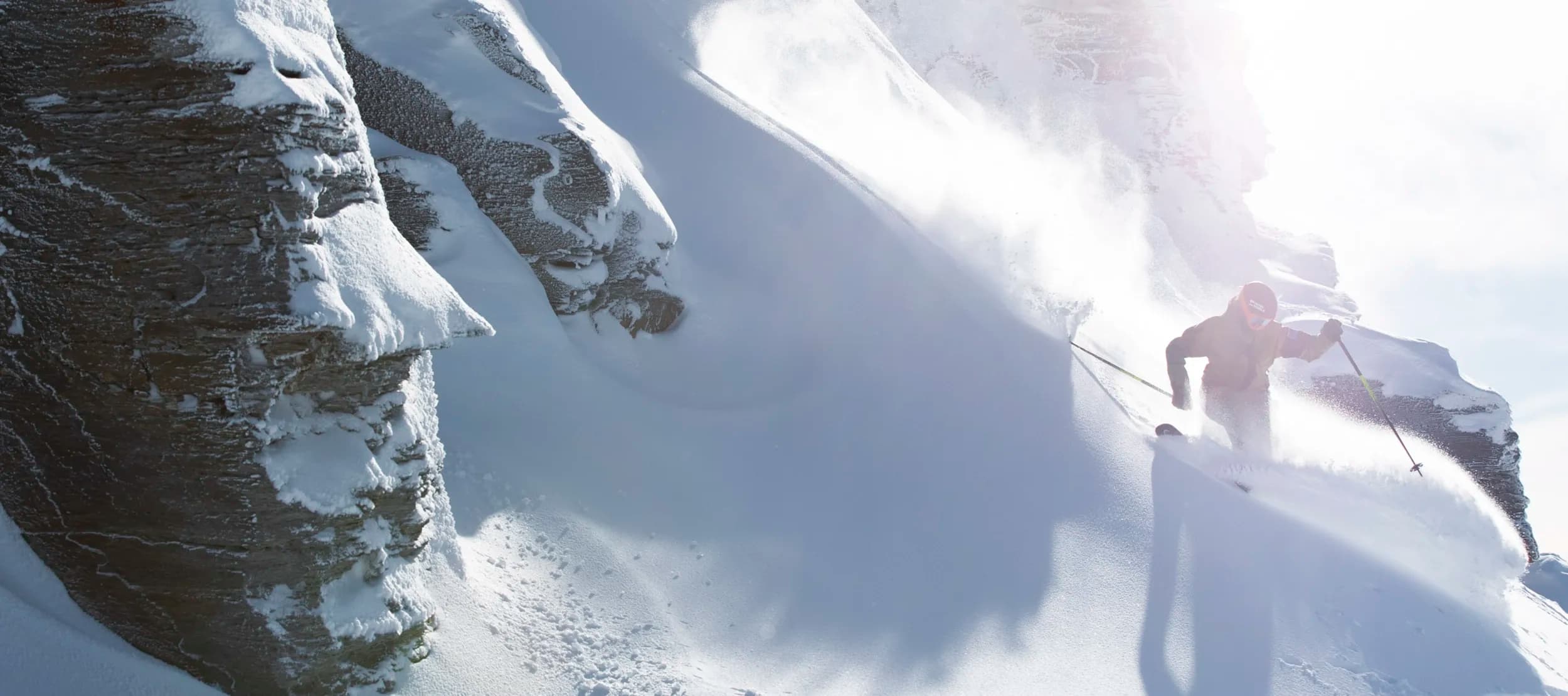 Skier carving down a steep, snow-covered mountain slope at Treble Cone Ski Field. Sunlight is illuminating the scene and casting shadows on the epic mountain landscape