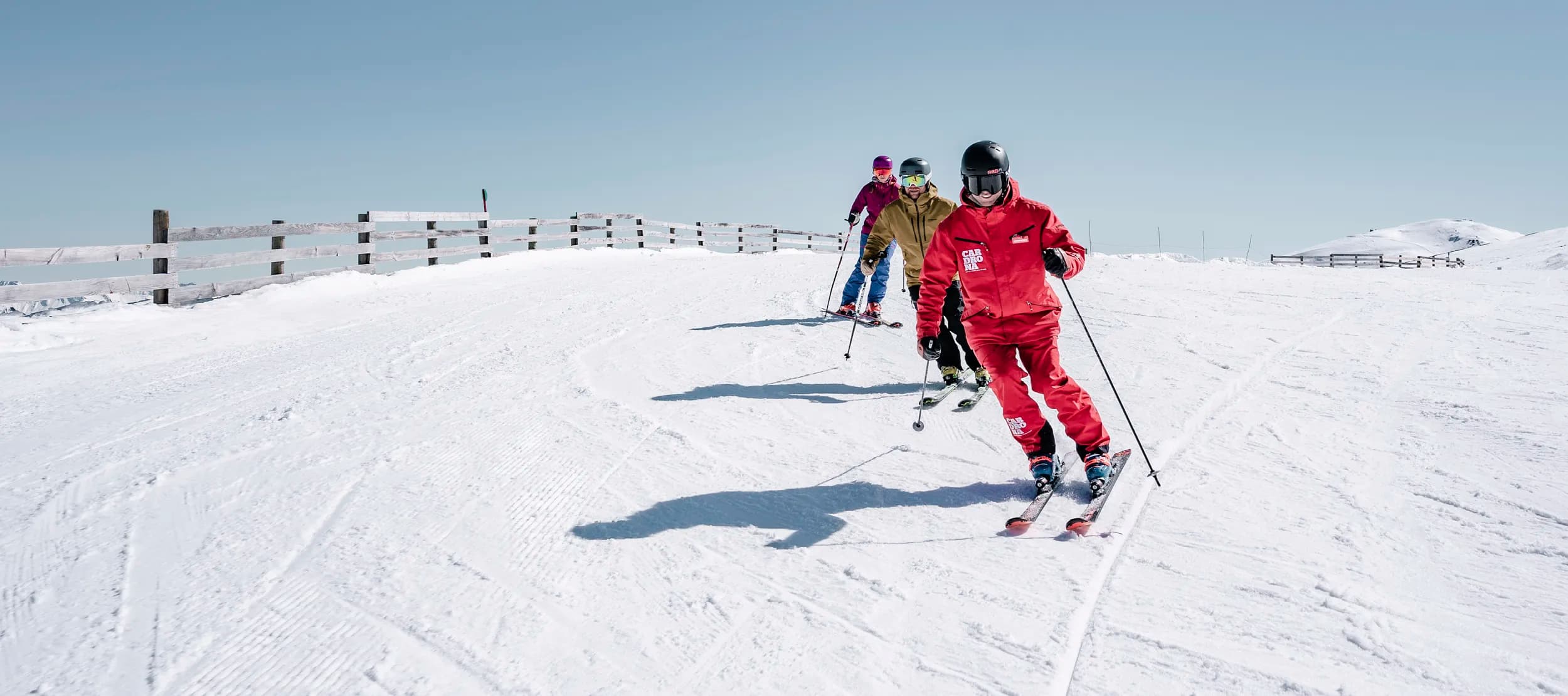 Three skiers on a snow-covered slope, following one another in a line. The skier in the front is dressed in a bright red outfit, while the two behind are in more muted colors. They are all wearing helmets and skiing under a clear blue sky, with a wooden fence lining the left side of the slope.