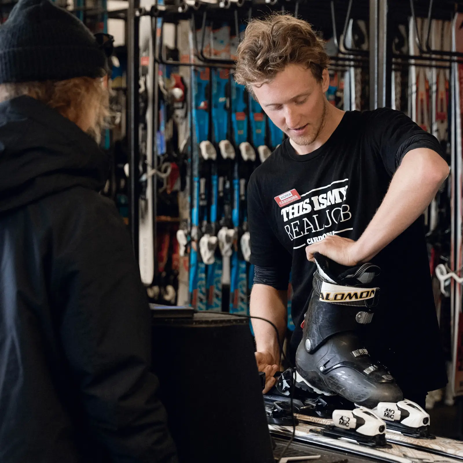 A rental technician at Cardrona Alpine Resort fits a ski boot for a guest in the rental shop, surrounded by racks of skis and snowboards.
