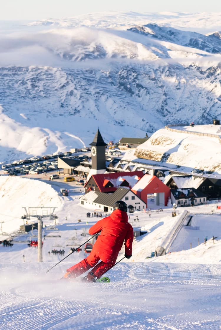 A skier dressed in red descends a freshly groomed slope at Cardrona, with the village, gondola, and chairlift visible in the background, all set against the snow-covered mountains.