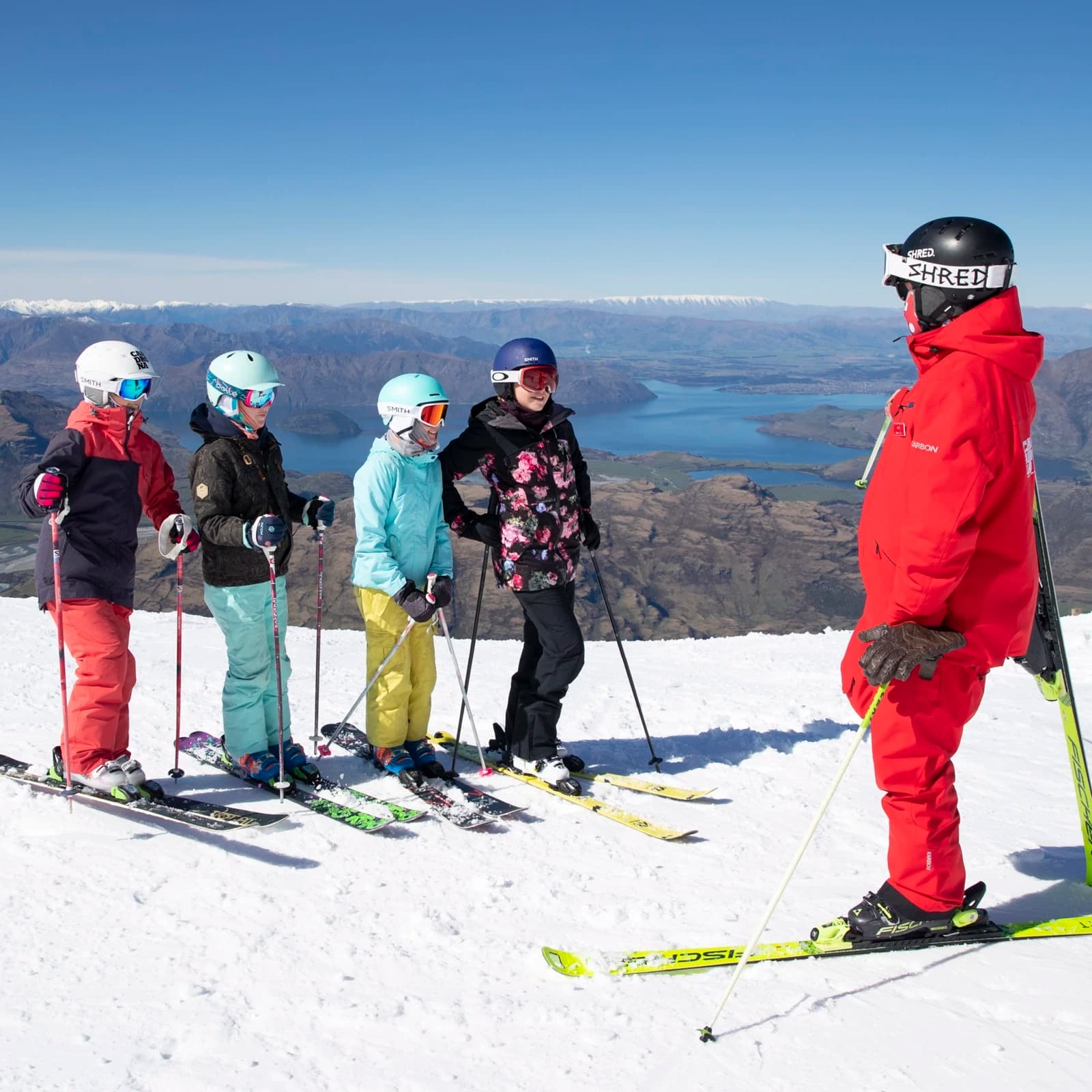A group of five skiers on a snowy mountain slope, posing with their skis. The group, comprising four children and one adult instructor, are all geared up with helmets and ski poles. They are dressed in colorful ski jackets and pants.